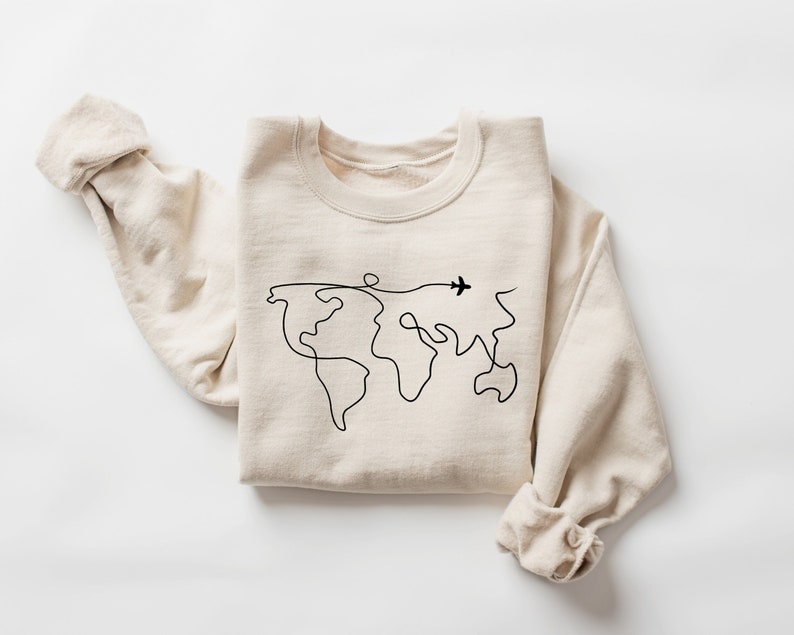 Op de afbeelding: Een beige sweatshirt met een zwarte lijntekening van de wereld en een klein vliegtuig dat erover vliegt.