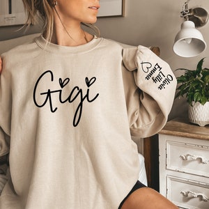 May include: A beige sweatshirt with the word "Gigi" printed on the front in black cursive lettering with two small hearts on either side. The left sleeve has the names "Oliver", "Lily", and "Emma" printed vertically with a heart at the top.