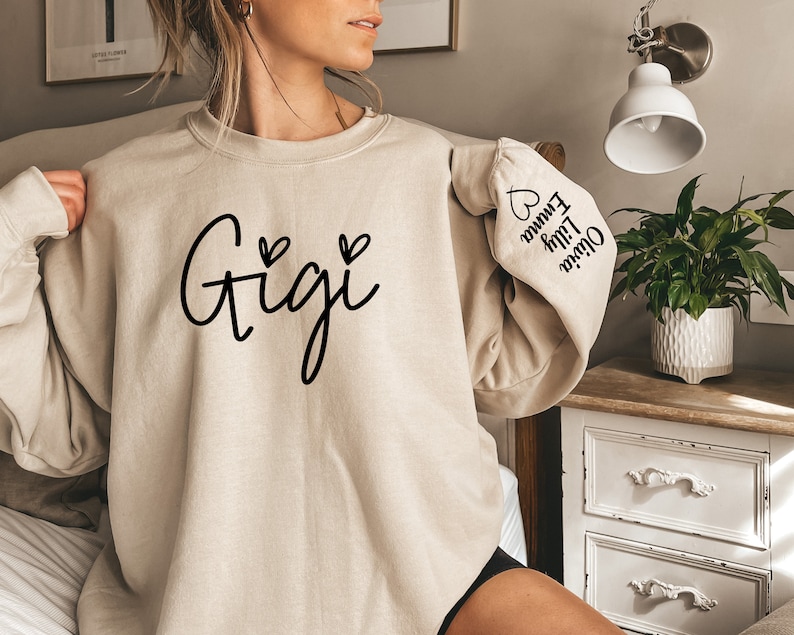 May include: A beige sweatshirt with the word "Gigi" printed on the front in black cursive lettering with two small hearts on either side. The left sleeve has the names "Oliver", "Lily", and "Emma" printed vertically with a heart at the top.