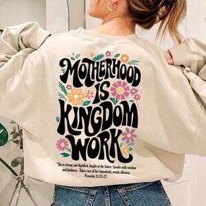 May include: A cream-colored sweatshirt with a graphic print that reads "Motherhood is Kingdom Work" in black with colorful flowers surrounding the text. The quote "She is strong and dignified, laughs at the future. Speaks with wisdom and kindness. Takes care of her household, avoids idleness. Proverbs 31:25-27" is printed below the graphic.