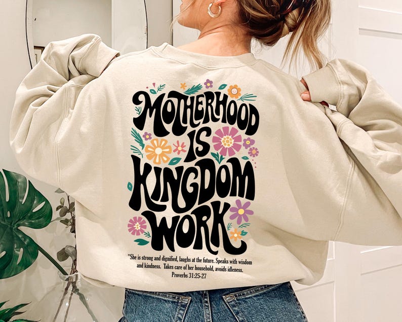May include: A cream-colored sweatshirt with a graphic print that reads "Motherhood is Kingdom Work" in black with colorful flowers surrounding the text. The quote "She is strong and dignified, laughs at the future. Speaks with wisdom and kindness. Takes care of her household, avoids idleness. Proverbs 31:25-27" is printed below the graphic.