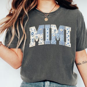 K&ouml;nnte beinhalten: Dunkelgraues T-Shirt mit dem Wort "MIMI" in einem Patchwork-Design. Die Buchstaben sind in blau-wei&szlig;en Blumen- und Vichy-Mustern gehalten. Das Shirt hat kurze &Auml;rmel und einen Rundhalsausschnitt.