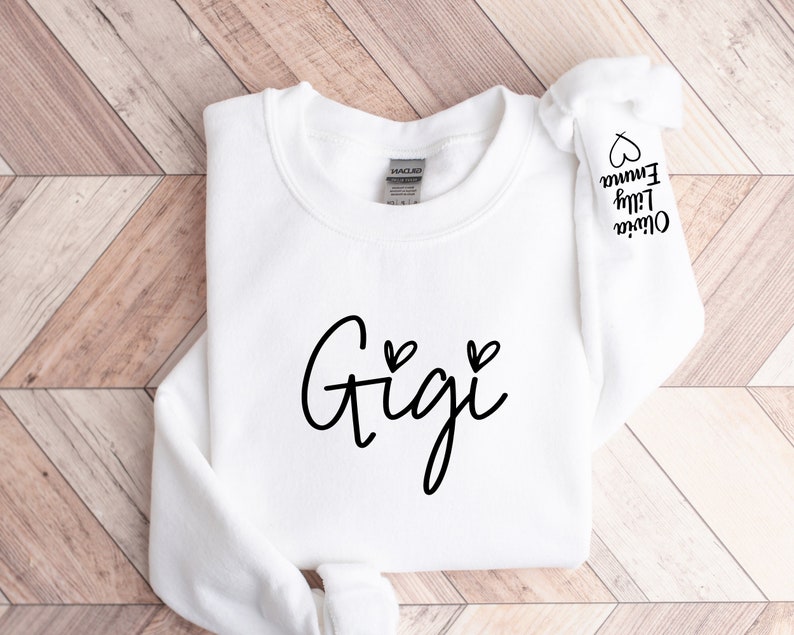 May include: White sweatshirt with the word "Gigi" written in black cursive with two small hearts above the word. The sleeve has the names "Oliver", "Lily", and "Emma" written in black cursive with a heart next to each name.
