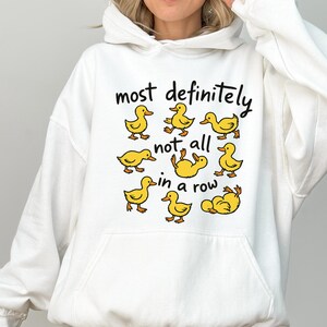May include: White hooded sweatshirt with a graphic of yellow cartoon ducks and the text "most definitely not all in a row". The ducks are in various poses. The sweatshirt has a front pocket and a drawstring hood.