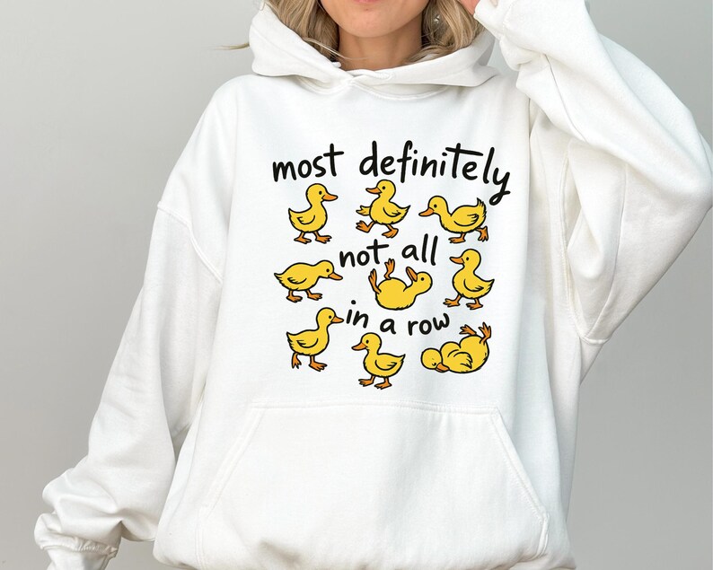 May include: White hooded sweatshirt with a graphic of yellow cartoon ducks and the text "most definitely not all in a row". The ducks are in various poses. The sweatshirt has a front pocket and a drawstring hood.