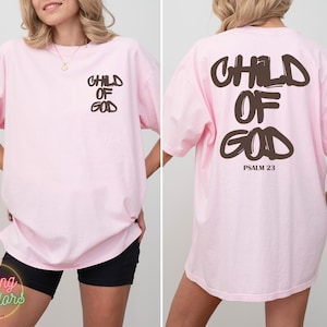 May include: Light pink oversized t-shirt with the words "CHILD OF GOD" printed in brown on the front and back. The back also includes "PSALM 23" in brown. The shirt is a comfortable, casual style.