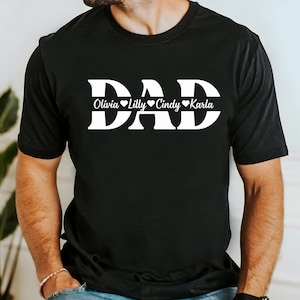 May include: Black t-shirt with the word "DAD" in large white letters. Below the word "DAD" is a white line with the names "Olivia", "Lilly", "Cindy", and "Karla" written in cursive with a heart symbol after each name.