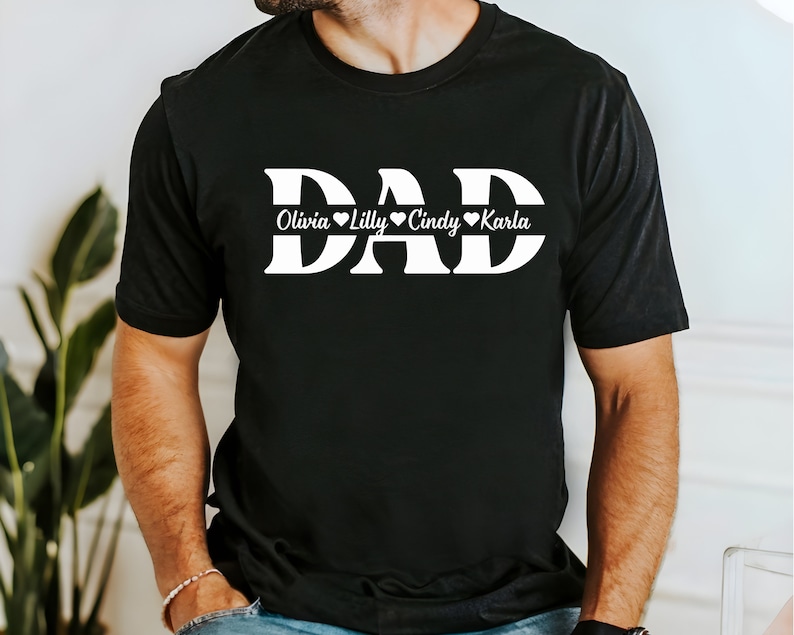 May include: Black t-shirt with the word "DAD" in large white letters. Below the word "DAD" is a white line with the names "Olivia", "Lilly", "Cindy", and "Karla" written in cursive with a heart symbol after each name.