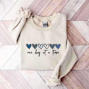 May include: A beige sweatshirt with a row of blue and white hearts and the text "one day at a time" printed on the front.