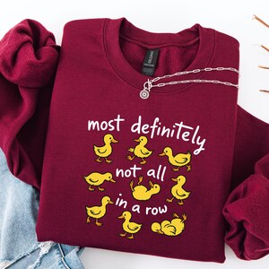 May include: Burgundy crewneck sweatshirt with the phrase "most definitely not all in a row" and yellow duck illustrations. A silver necklace with a circular pendant is draped across the sweatshirt. The sweatshirt is folded on top of a pair of blue jeans.