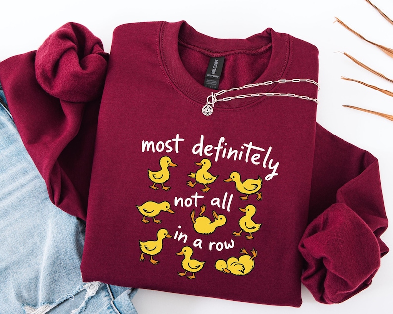 May include: Burgundy crewneck sweatshirt with the phrase "most definitely not all in a row" and yellow duck illustrations. A silver necklace with a circular pendant is draped across the sweatshirt. The sweatshirt is folded on top of a pair of blue jeans.