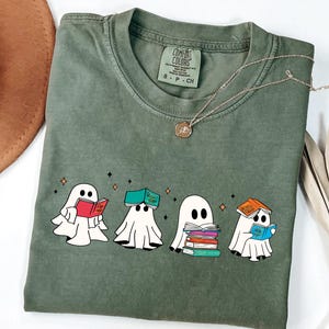 May include: Olive green Comfort Colors t-shirt featuring a graphic of four cartoon ghosts reading books. Each ghost is a different color, and the books are red, green, white, and blue. A gold necklace is draped across the shirt.