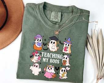 May include: Olive green t-shirt with a crew neck and the phrase "Teaching My Boos" printed in black. The shirt features a graphic of cartoon ghosts, each with unique accessories, including a graduation cap, witch hat, and coffee cup.