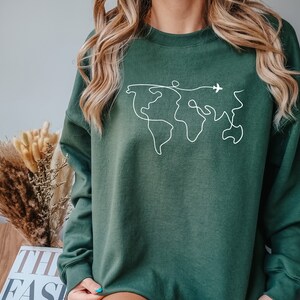 Op de afbeelding: Een donkergroen sweatshirt met een witte omtrek van een wereldkaart en een klein vliegtuig dat erover vliegt.