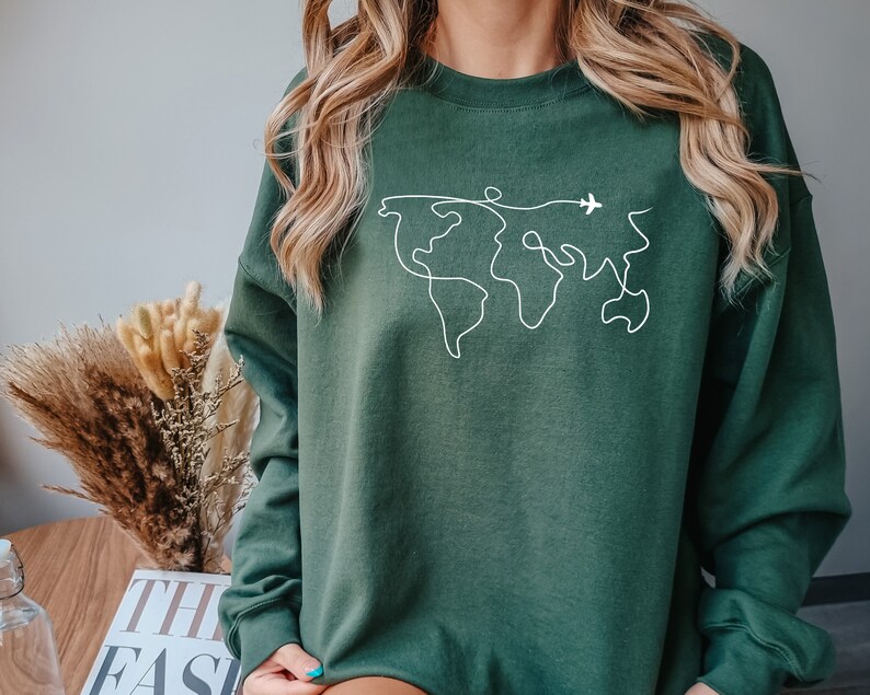 Op de afbeelding: Een donkergroen sweatshirt met een witte omtrek van een wereldkaart en een klein vliegtuig dat erover vliegt.