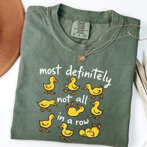 May include: Green Comfort Colors t-shirt with the phrase "most definitely not all in a row" and yellow duck illustrations. The shirt has a crew neck and short sleeves. A gold necklace is draped across the shirt.