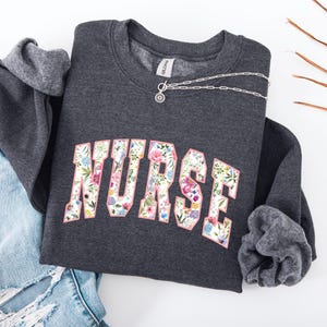 May include: A dark gray sweatshirt with a floral print of the word "NURSE" in pink.