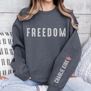 May include: Dark gray sweatshirt with the word "FREEDOM" in distressed white lettering across the chest. The right sleeve features the text "CHARLIE KIRK" in white, with a small American flag graphic.