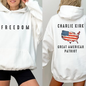 May include: White hooded sweatshirt with the word "FREEDOM" printed in black on the front. The back of the sweatshirt features the text "CHARLIE KIRK GREAT AMERICAN PATRIOT" with an American flag design in the shape of the United States.