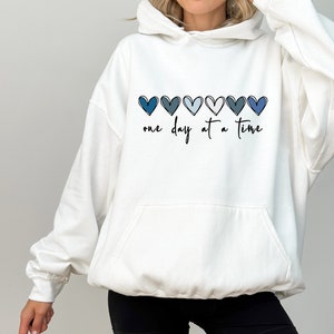 May include: White hooded sweatshirt with a blue ombre heart design and the text "one day at a time".