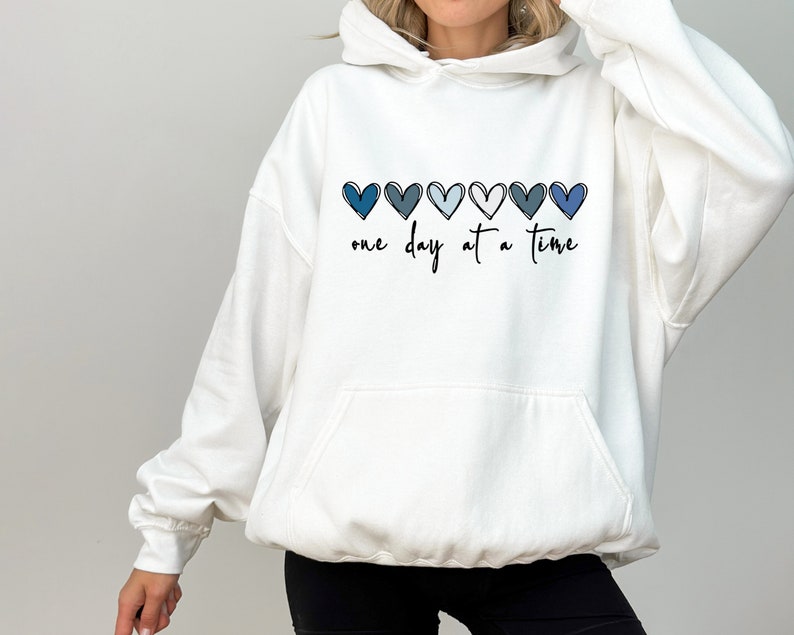May include: White hooded sweatshirt with a blue ombre heart design and the text "one day at a time".