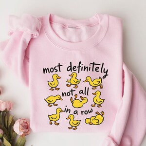 May include: A light pink sweatshirt with the phrase "most definitely not all in a row" in black script. The sweatshirt features cartoon yellow ducklings in various poses. The sleeves are folded, and the background is white.