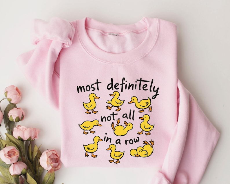 May include: A light pink sweatshirt with the phrase "most definitely not all in a row" in black script. The sweatshirt features cartoon yellow ducklings in various poses. The sleeves are folded, and the background is white.