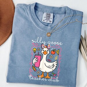 May include: A blue Comfort Colors t-shirt with the words "silly goose teacher club" and a cartoon goose holding a pink drink. The goose has a bow and is surrounded by school-themed graphics. A gold necklace is draped across the shirt.