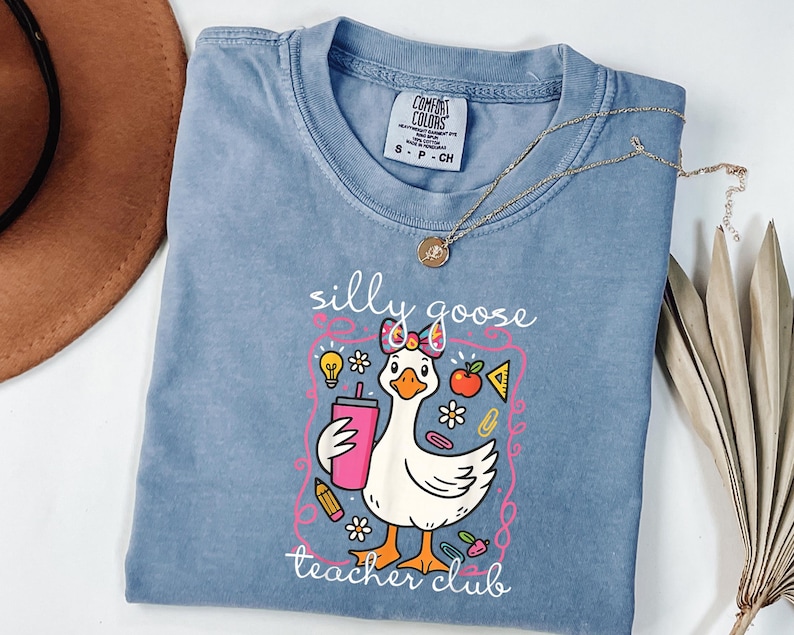 May include: A blue Comfort Colors t-shirt with the words "silly goose teacher club" and a cartoon goose holding a pink drink. The goose has a bow and is surrounded by school-themed graphics. A gold necklace is draped across the shirt.