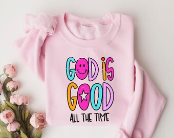 May include: A light pink sweatshirt with the words "GOD IS GOOD ALL THE TIME" in colorful, distressed lettering. The design includes a smiley face and a star. The sweatshirt is folded on a white surface with pink flowers.