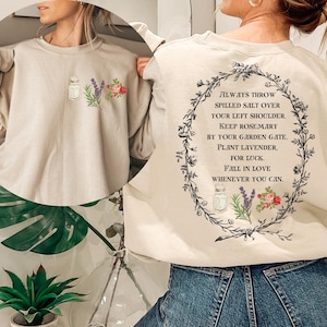 May include: A beige sweatshirt with a floral design and the text "ALWAYS THROW SPILLED SALT OVER YOUR LEFT SHOULDER. KEEP ROSEMARY BY YOUR GARDEN GATE. PLANT LAVENDER. FOR LUCK. FALL IN LOVE WHENEVER YOU CAN."