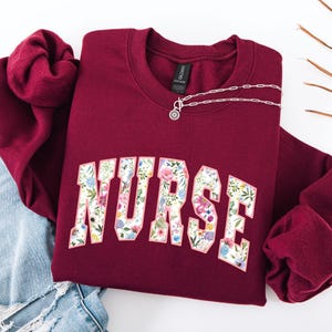 May include: Burgundy sweatshirt with a floral print of the word "NURSE" on the front.