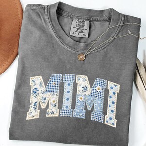 K&ouml;nnte beinhalten: Graues Comfort Colors T-Shirt mit dem Namen "MIMI" in einem Patchwork-Design aus blauem Blumen-, Vichy- und Tupfenstoffen. Eine silberne Halskette mit Anh&auml;nger liegt auf dem Shirt. Ein brauner Hut ist im Hintergrund zu sehen.