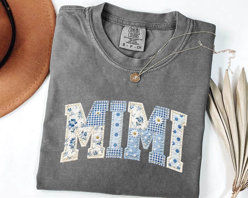 K&ouml;nnte beinhalten: Graues Comfort Colors T-Shirt mit dem Namen "MIMI" in einem Patchwork-Design aus blauem Blumen-, Vichy- und Tupfenstoffen. Eine silberne Halskette mit Anh&auml;nger liegt auf dem Shirt. Ein brauner Hut ist im Hintergrund zu sehen.