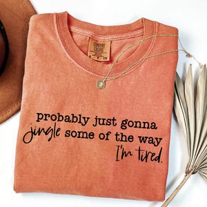 May include: Coral-coloured Comfort Colors t-shirt with the phrase "probably just gonna jingle some of the way I'm tired" in black script. A gold necklace with a pendant rests on the shirt. A brown hat and dried palm leaves are in the background.