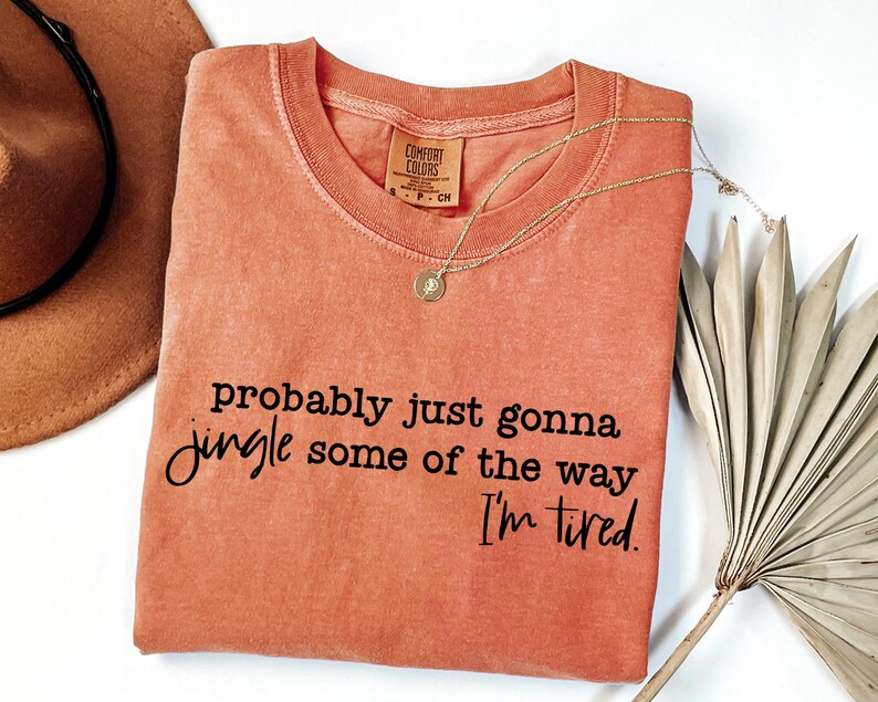 May include: Coral-coloured Comfort Colors t-shirt with the phrase "probably just gonna jingle some of the way I'm tired" in black script. A gold necklace with a pendant rests on the shirt. A brown hat and dried palm leaves are in the background.