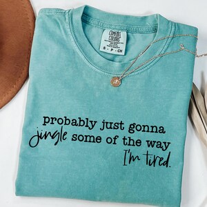 May include: A turquoise Comfort Colors t-shirt with the text "probably just gonna jingle some of the way I'm tired." A gold necklace with a pendant is draped across the shirt. A brown hat and dried plant are in the background.