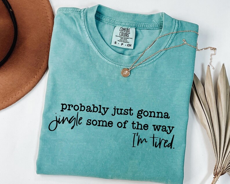 May include: A turquoise Comfort Colors t-shirt with the text "probably just gonna jingle some of the way I'm tired." A gold necklace with a pendant is draped across the shirt. A brown hat and dried plant are in the background.