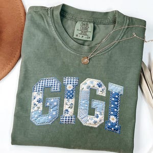 May include: Olive green Comfort Colors t-shirt with the name "GIGI" appliqued in blue and white floral and gingham fabric. A gold necklace with a pendant rests on the shirt. A brown hat and dried palm leaves are in the background.