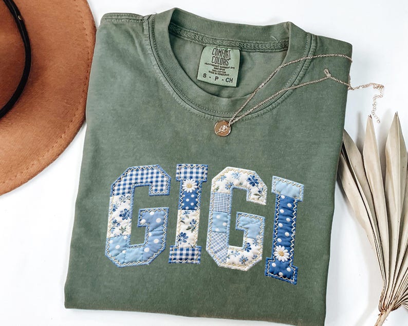 May include: Olive green Comfort Colors t-shirt with the name "GIGI" appliqued in blue and white floral and gingham fabric. A gold necklace with a pendant rests on the shirt. A brown hat and dried palm leaves are in the background.