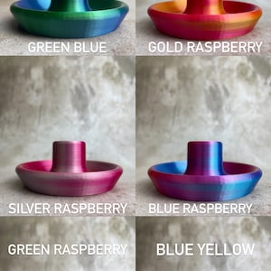 May include: A selection of 3D-printed ashtrays in various colour combinations. Each ashtray has a circular base with a central post. The colour schemes include green and blue, gold and raspberry, silver and raspberry, and blue and yellow.