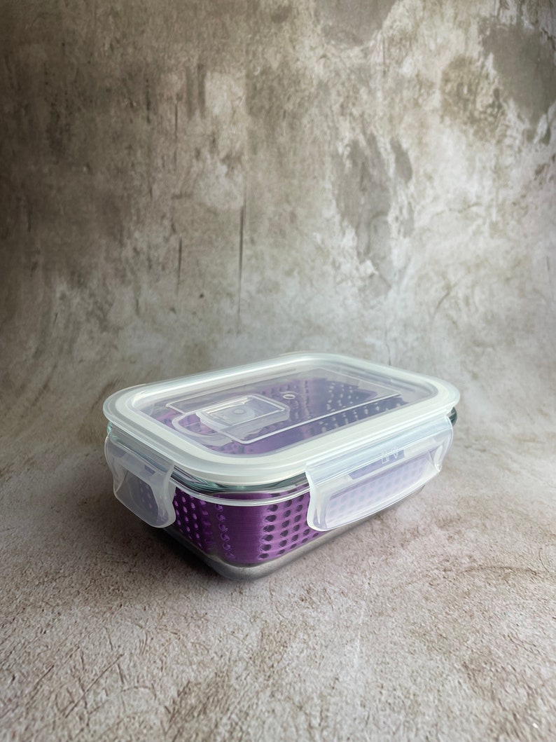 May include: A rectangular glass food storage container with a clear lid and purple interior. The lid has white plastic clips on each side. The container is designed for food storage and is suitable for kitchen use.