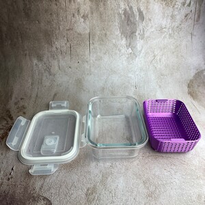 May include: A clear glass rectangular food storage container with a white lid and purple plastic insert. The lid is open, revealing a white seal. The container is designed for food storage.