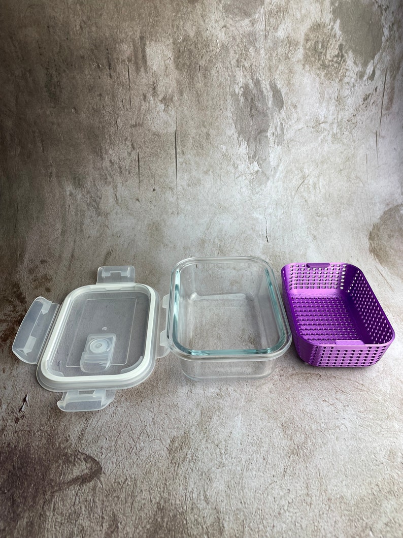 May include: A clear glass rectangular food storage container with a white lid and purple plastic insert. The lid is open, revealing a white seal. The container is designed for food storage.