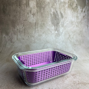 May include: A rectangular glass food container with a removable, perforated purple insert. The container has a clear glass exterior. The purple insert is designed with numerous small holes. The container is set against a neutral background.