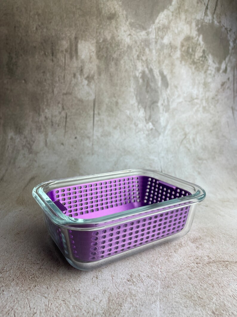 May include: A rectangular glass food container with a removable, perforated purple insert. The container has a clear glass exterior. The purple insert is designed with numerous small holes. The container is set against a neutral background.