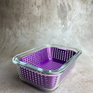 May include: A rectangular glass container with a vibrant purple, perforated insert. The clear glass sides and top allow a view of the interior. The container sits on a neutral-toned surface.
