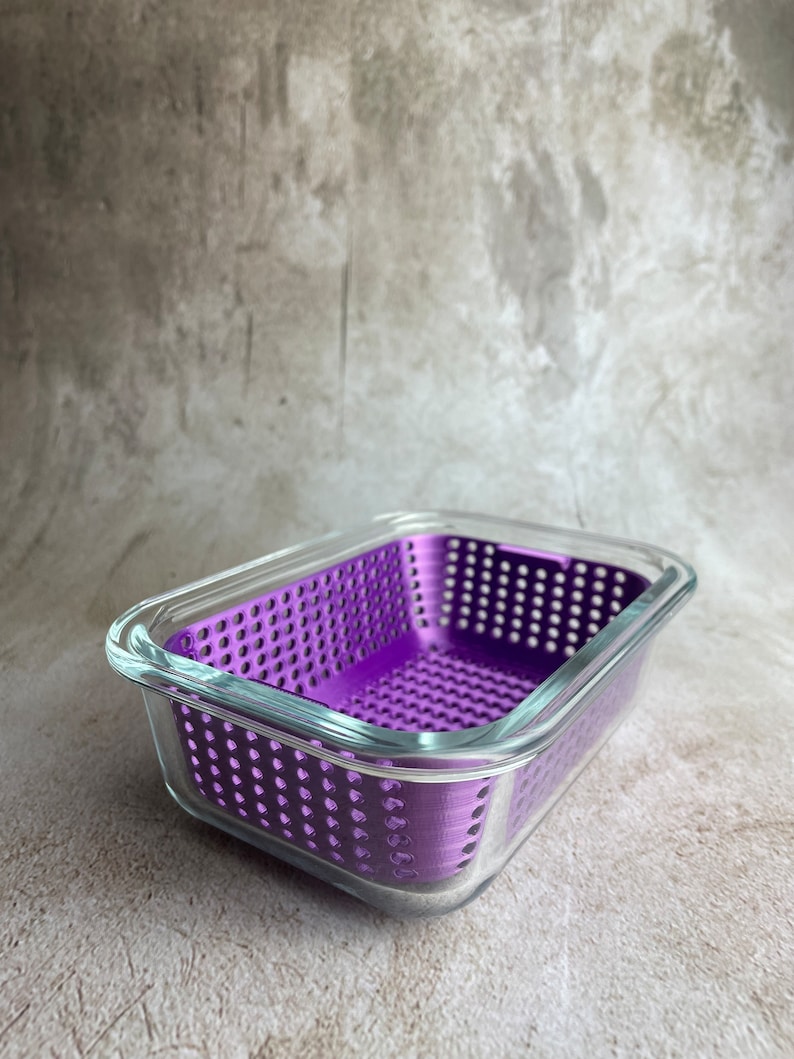 May include: A rectangular glass container with a vibrant purple, perforated insert. The clear glass sides and top allow a view of the interior. The container sits on a neutral-toned surface.