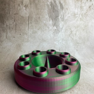 May include: A 3D-printed, circular object with a central cone and ten evenly spaced cylindrical holes. The object is a blend of green and magenta colors, with a textured appearance. The design suggests a holder or organizer.