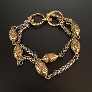 May include: A gold and silver-toned bracelet with an ornate design. The bracelet features gold-colored oval beads with detailed patterns, connected by silver-colored chain links. The clasp is also gold-colored, with a decorative hook closure.